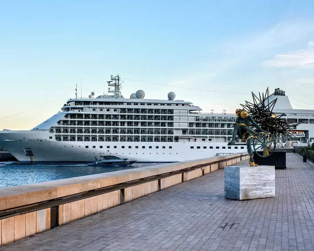 A large, white cruise ship is docked at a port. The ship has multiple decks and a distinctive design. In the foreground, there is a paved walkway with a sculpture on a pedestal. The sky is blue and clear.