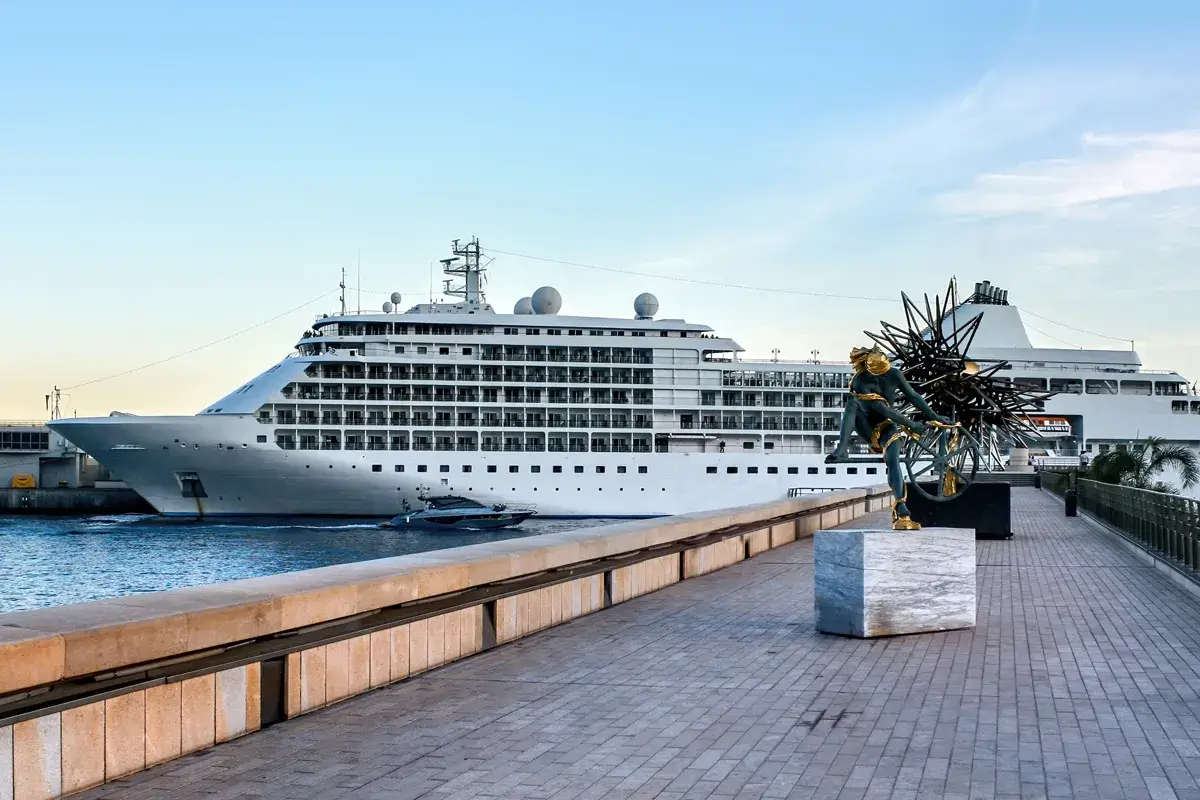 A large, white cruise ship is docked at a port. The ship has multiple decks and a distinctive design. In the foreground, there is a paved walkway with a sculpture on a pedestal. The sky is blue and clear.